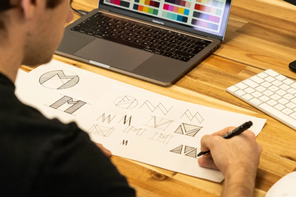 Persona bosquejando propuestas de logo junto a una computadora, imagen representativa del diseño de identidad de marca