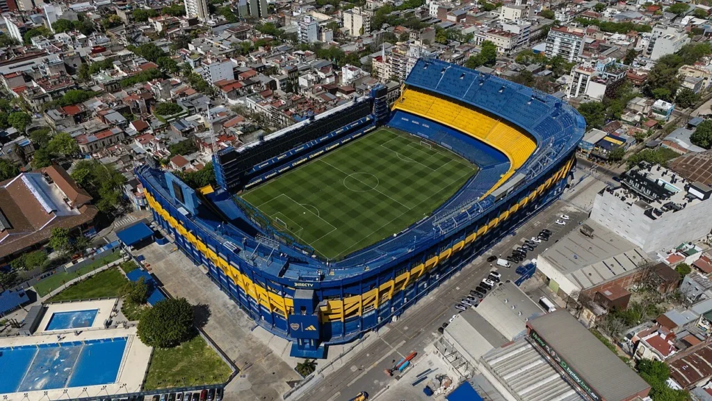 Vista aérea de La Bombonera, con tribunas azules y amarillas y cancha de fútbol, rodeada por edificios del barrio de La Boca.