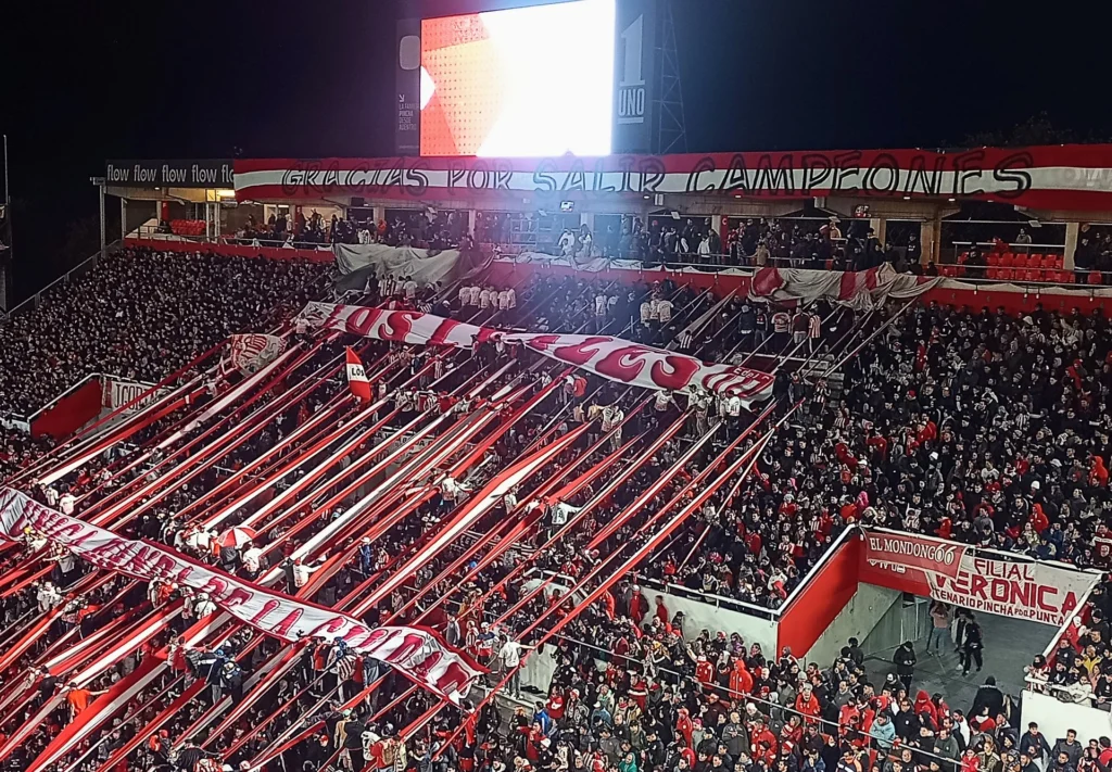 Tribuna de la hinchada de Estudiantes de La Plata con banderas y tiras rojas y blancas en el estadio UNO.